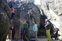 Mehrere Personen zwischen Sandsteinblöcken; eine Kletterin an einer Wand, ca. 2&nbsp;m über dem Boden; ein Mann hat beide Arme in Richtung der Kletterin ausgebreitet