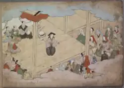 Okuni steht auf einer Bühne, vor der mehrere Zuschauer sitzen. Sie trägt einen schwarzen Kegelhut, ein beigenes Oberteil und eine weite, schwarze Hose mit weißem Pflanzenmuster. In der linken Hand hält sie einen Gong, in der rechten einen schwarzen Gongschlägel. Hinter ihr sitzen mehrere als Priester verkleidete Schauspieler. Sie tragen einen Chonmage, kahl rasiertes Haupthaar plus Pferdeschwanz. Die Priester tragen verschiedenfarbige Gewänder, einer von ihnen schlägt eine kleine Trommel.