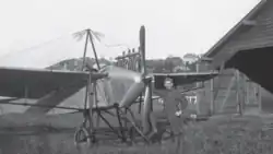 Schwarzweißfotografie eines frühen Propellerflugzeugs. Es steht neben einem Schuppen. Vor dem Propeller steht eine Person.