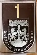 Plakette für Pirna in Form eines Wappenschilds, mit goldfarbener Hausnummer und dem Pirnaer Wappen darunter (vermutlich Aluminiumguss).