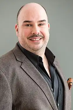 Robert Joseph Bartl beim Bayerischen Fernsehen Filmbrunch 2016, ein Mann mit Glatze und kurzem Bart, lächelt freundlich. Er trägt einen braunen Blazer mit einem dunklen Hemd und einer schwarzen Weste darunter. Der Blazer hat ein klassisches Revers, und in der Brusttasche steckt ein orangefarbenes Einstecktuch. Der Hintergrund ist schlicht und grau.