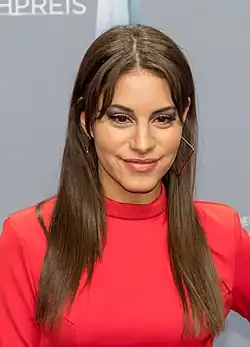 Almila Bagriacik beim Deutscher Fernsehpreis 2018. Almila Bagriacik mit langen, glatten, braunen Haaren, die ein rotes Oberteil trägt. Sie steht vor einem grauen Hintergrund, auf dem teilweise der Schriftzug „PREIS“ sichtbar ist. Die Person trägt auffällige, geometrische Ohrringe, die einen modernen und stilvollen Eindruck vermitteln.