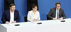 Drei Personen sitzen an einem Tisch und unterschreiben Dokumente. Sie wirken konzentriert und professionell. Der Hintergrund ist eine schlichte blaue Wand.