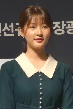 Kim Sae-ron (2018), die ein dunkelgrünes Kleid mit einem weißen Kragen trägt. Das Kleid ist mit einer Reihe dekorativer Knöpfe verziert, die vorne verlaufen. Der Hintergrund ist schwarz, und darauf befindet sich weiße koreanische Schrift. Die Schrift ist nur teilweise sichtbar und scheint Teil eines größeren Schildes oder Banners zu sein.