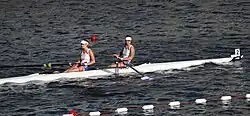 Das Bild zeigt das Boot von der Seite. Beide Ruderinnen tragen ein weißes Trikot und eine dunkelblaue Hose.