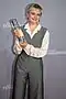 Lena Urzendowsky beim Deutschen Fernsehpreis 2019 in der Düsseldorfer Rheinterrasse hält den ihr verliehenen Preis in der Hand, trägt einen grauen Overall und ein weißes Hemd, lächelt in die Kamera.Der Fernsehpreis ist eine transparente, rechteckige Trophäe mit einer sichtbaren Inschrift darauf. Sie trägt ein graues ärmelloses Overall-Outfit, unter dem ein weißes langärmliges Hemd zu sehen ist. Dazu trägt sie rote Ohrringe, die einen farblichen Akzent setzen. Der Hintergrund des Bildes ist grau und weist die Aufschrift „DER DEUTSCHE FERNSEHPREIS“ auf.