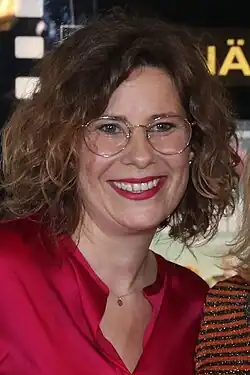 Nicole Kersten lächelt freundlich, mit schulterlangem, lockigem Haar, die eine rote Bluse trägt. Sie hat eine kleine, runde Anhänger-Halskette um den Hals. Im Hintergrund ist ein schwarzes Schild mit gelber Schrift sichtbar, auf dem „Ä“ geschrieben steht. Rechts im Bild ist eine weitere Person teilweise zu sehen, die ein gestreiftes, glitzerndes Oberteil trägt. Das Foto entstand bei der Premiere des Fernsehfilmes „Klassentreffen“ 2019.