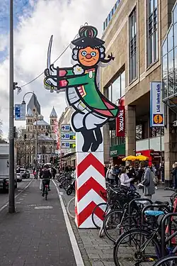 Straßenszene in Köln mit einer großen bunten Karnevalsfigur auf einem rot-weiß gestreiften Sockel am Straßenrand. Die Figur stellt eine stilisierte, lächelnde Person in grün-rotem Uniformkostüm mit Dreispitz und Degen dar – typisch für den Kölner Karneval. Im Hintergrund sieht man Passanten, Fahrradständer, Geschäftsschilder (u. a. Lidl, TK Maxx) und weiter hinten die romanische Kirche Groß St. Martin mit ihren markanten Türmen. Es ist tagsüber, der Himmel ist bewölkt, und es herrscht reger Fuß- und Radverkehr.