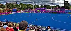 Farbfoto mit einer Totalen von der Tribüne über den blauen Kunstrasen auf die Gegentribüne