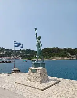 Die Statue aus korrodierter Bronze zeigt einen jungen Mann mit einer Fackel in der erhobenen Hand auf dem Bug eines Schiffes stehend. Die Statue steht auf einem Steinsockel. Daneben weht eine griechische Flagge. Im Hintergrund ist der Segelhafen von Gaios zu sehen.