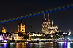 nächtliches Rheinpanorama Kölns (Dom, Groß-St. Martin, Rathaus, Schiffe, Fluss) mit farbigen Strahlen in Regenbogenfarben am Himmel