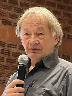 Thomas Bock mit einem Mikrofon in der rechten Hand vor einer Ziegelwand