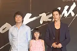 Kim Sae-ron (2010) in einem pinkfarbenen Kleid, mit Regisseur Lee Jeong-beom trägt ein hellblaues Hemd und Won Bin trägt einen dunklen Blazer über einem schwarzen Hemd bei einer Veranstaltung zum Film The Man from Nowhere.Die dreier Gruppe steht vor einem dunklen Hintergrund mit weißer, schwungvoller Textgestaltung. Die Textelemente wirken wie kräftige Pinselstriche und sind in koreanischer Schrift.
