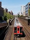 Großstadtszenerie: Rechts und links mehrstöckige Gebäude, in der Mitte eine viergleisige Eisenbahnstrecke, rechts mit gemauerter Stützwand, links mit Brüstung. Auf dem zweiten Gleis von links fährt ein rot-schwarz lackierter Triebwagenzug auf die Kameraposition zu, die sich auf einer Querstraße oberhalb der Gleise befindet und den Blick schräg aufs mit silbrigen Riffelblechen verkleidete Dach des Zuges fallen lässt.