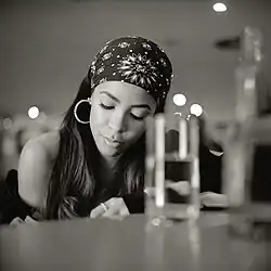 Aaliyah schwarz-weiße Fotografie, die an einem Tisch oder an Klavier sitzt. Sie trägt ein Bandana mit floralem Muster und große Creolen-Ohrringe. Das lange Haar der Person ist sichtbar, und sie scheint ein ärmelloses Oberteil zu tragen. Auf dem Tisch vor der Person befinden sich mehrere Gläser und Flaschen, die ebenfalls etwas unscharf sind. Im Hintergrund ist eine verschwommene Umgebung mit einigen Lichtquellen sichtbar, die auf einen schwach beleuchteten Raum hinweist, möglicherweise ein Restaurant oder eine Bar.