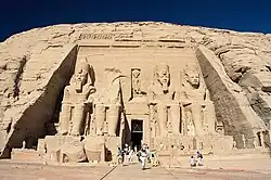 Frontale Farbfotografie eines großen Felsens, in deren Mitte vier riesige, sitzende Steinpharaonen gemeißelt sind. Oberkörper und Kopf des zweiten Pharaos liegen zertrümmert vor seinen Füßen. Zwischen ihnen ist ein dunkler Eingang, durch den viele Menschen gehen. Über dem Eingang ist eine kleine Figur in den flach geschliffenen Felsenteil eingemeißelt.