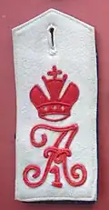 Fotografie einer weißen Schulterklappe mit aufgesticktem roten Schnörkelbuchstaben "A", darüber eine Zarenkrone