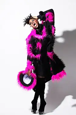 Porträtfoto von Alex Newell, der vor einer weißen Wand steht. Er hat das linke Auge geschlossen und das rechte geöffnet. Er trägt rosa Lippenstift, blauen Lidschatten, Wimperntusche, ein schwarzes Oberteil mit schwarzen und pinken Plüschstellen, schwarze Strümpfe und schwarze High Heels. In der rechten Hand hält er einen kleinen schwarzen Stoffring mit pinker Plüschumrandung. Die linke Hand hält sich Newell an den Kopf. Seine schwarzen Haare trägt er als nach oben abstehende, gekräuselte Frisur.