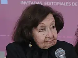 Amparo Dávila sitzt vor einem Tisch, wo ihre Name steht und ihrem Buch Árboles petrificados. Eine Frau hält das Mikrofon, damit sie sprechen kann, weil sie bereits sehr alt ist.