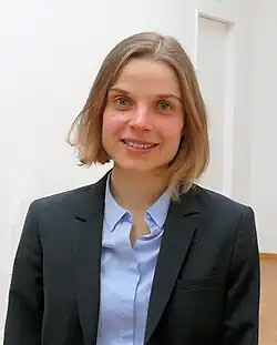 Andrea Ablasser beim Empfang des Ludwig Darmstaedter-Nachwuchspreises 2014. Sie trägt einen schwarzem-dunklen Blazer über einem hellblauen Hemd mit Kragen. Ihr Haar ist kurz und hellbraun, und sie hat einen professionellen und eleganten Look. Der Hintergrund des Bildes zeigt einen Innenraum mit weißen Wänden und einem Holzboden, was eine schlichte und formelle Atmosphäre vermittelt.