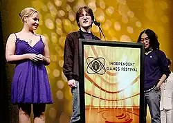 Zwei Männer stehen hinter einem Podium mit der Aufschrift Independent Games Festival, während eine Frau in einem blauen Kleid links steht.