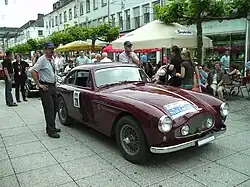 Hier ist ein weinrotes Auto abgebildet, bei Tageslicht auf einer Straße mit vielen Menschen in der Nähe.
