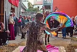 Im Innenhof eines alten Fachwerkhauses stehen gut dreißig Menschen versammelt und blicken auf eine Drag Queen in der Bildmitte und einen Mann im Vordergrund, der einen silbernen Paillettenanzug trägt und kunstvoll eine kleine Regenbogenflagge durch die Luft schwingt.