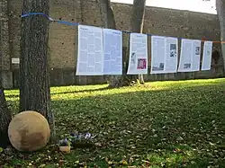 Biografietafeln sind auf einer Schnur zwischen Bäumen hängend zu sehen. Links im Bild liegt die gedenkkugel, im Hintergrund ist die Außenmauer des Lagers zu sehen.