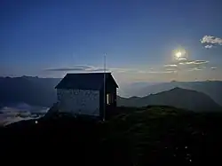 Eine Biwackhütte, die mit Holz verkleidet ist, vor einem Bergpanorama bei Nacht, wobei der Mond hell auf die Szenerie scheint.