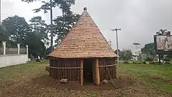 Rundhaus mit spitzem Dach, die niedrigen Wände sind aus senkrechten Rundhölzern. Dem Fotografen zugewandt ist eine offene Türe.