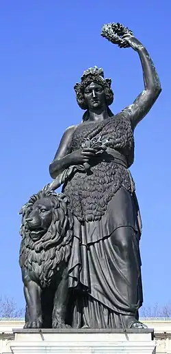 Farbige Untersicht auf eine weibliche Figur in griechischem Stil mit einem Löwen an der linken Seite. Die Figur drückt mit ihrer rechten Hand ein Schwert an ihre Brust und hält mit ihrer linken Hand einen Lorbeerkranz über ihr Haupt.