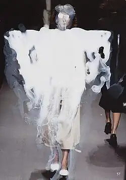 Ein Model läuft in einem Kleidungsstück über den Laufsteg, das mit weißer Farbe grob überstrichen ist, auch das Gesicht ist mit weißer Farbe verfremdet, was einen einen surrealen Effekt erzeugt.