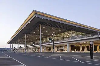 Seitliche Farbfotografie von einem zweigeschossigen Gebäude mit Stützpfeilern im Erdgeschoss und breiter Glasfront in der obersten Etage. Das weit überragende Dach wird von großen Pfeilern gestützt und hat Spots.