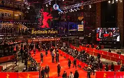 Farbige Obersicht auf ein Glasgebäude mit einem roten Teppich und vielen Presseleuten im Vordergrund. Rechts zeigt ein Bildschirm ankommende Gäste. Viele Zuschauer sind im Gebäude mit der Aufschrift „Berlinale Palast. Theater am Potsdamer Platz“ und ein roter Berliner Bär als Logo. Auf der linken Seite innerhalb des Gebäudes hängen einige Porträts.