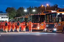Das Bild zeigt die frühen Morgenstunden der Müllabfuhr. Die Müllwerker:innen beginnen den Arbeitstag. Der Fuhrpark ist zu sehen.