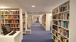 Blick in den langen Gang der PRIF-Bibliothek mit beidseitig hohen Bücherregalen und einem Computerarbeitsplatz links vorne.