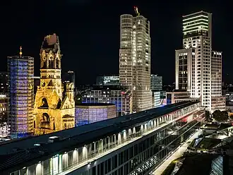 Nachtfotografie im Bereich um den Zoologischen Garten. Im Vordergrund steht ein langes modernes Gebäude. Dahinter ist eine Kirchenruine, neben der links ein achteckiger Turm und rechts ein achteckiges Gebäude stehen. Die Fronten bestehen aus blauen Kirchenfenstern. Rechts von der Kirche sind zwei Hochhäuser.