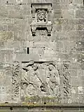 Relief auf der Westseite Oben ein angedeutetes Fenster mit Gesicht und der Überschrift: KRIEG Unten: Heldenjüngling im Kampf gegen einen Drachen und Befreiung einer Jungfrau.