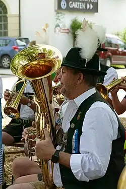 Foto des Oberkörpers eines Mannes, der in ein Blechblasinstrument bläst. Er trägt einen grünen Hut mit Federn. Im Hintergrund sind weitere Musiker zu sehen