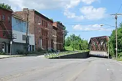 mehrere alte Backsteingebäude an der linken Seite einer Straße, die zu einer rostigen Eisenbahnbrücke hin abfällt
