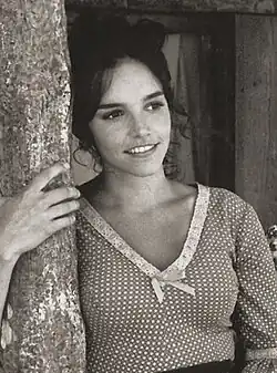 Brooke Adams mit dunklen, lockigen Haaren, lehnt an einem Baum und lächelt freundlich. Sie trägt eine gemusterte Bluse mit einem V-Ausschnitt, der mit Spitzenbesatz und einer kleinen Schleife in der Mitte versehen ist. Die linke Hand von ihr ruht auf dem Baumstamm. Im Hintergrund ist eine Holzstruktur zu sehen, möglicherweise ein Haus oder eine Hütte.