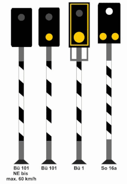 Vier Lichtsignale, die jeweils eine rechteckigen schwarzen Signalschirm besitzen und ein schwarz-weiß schräg gestreiftes Mastschild tragen. Von links nach rechts handelt es sich um die folgenden Signale: 1. Bü 101 (NE bis max. 60 km/h): Ein weiß blinkendes Licht; 2. Bü 101: Ein weiß blinkendes Licht über einem gelben Licht; 3. Bü 1: Der Mastschirm trägt einen gelben Rahmen, darin oben ein weiß blinkendes Licht, darunter eine gelbe Kreisscheibe; 4. So 16a: Ein weißes (dauerleuchtendes) Licht, darunter nebeneinander zwei gelbe Lichter.