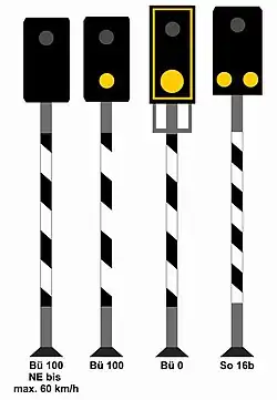 Vier Lichtsignale, die jeweils eine rechteckigen schwarzen Signalschirm besitzen und ein schwarz-weiß schräg gestreiftes Mastschild tragen. Von links nach rechts handelt es sich um die folgenden Signale: 1. Bü 100 (NE bis max. 60 km/h): Ein erloschenes Licht; 2. Bü 100: Ein erloschenes Licht über einem gelben Licht; 3. Bü 0: Der Mastschirm trägt einen gelben Rahmen, darin oben ein erloschenes Licht, darunter eine gelbe Kreisscheibe; 4. So 16b: Ein erloschenes Licht, darunter nebeneinander zwei gelbe Lichter.