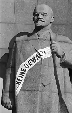 Schwarzweißfoto einer steinernen Statue von Lenin vor steinernem Hintergrund, über der Brust eine weiße Schärpe mit der Aufschrift „Keine Gewalt!“