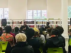 Candice Carty-Williams sitzt in einer Bibliothek vor einer Gruppe von Menschen. Sie hält ein Mikrofon in der Hand. Im Hintergrund sie die Bücherregale der Bibliothek zu sehen.