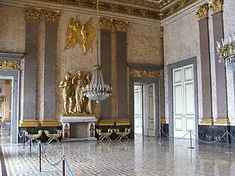 Farbfotografie eines Innenraums mit drei goldenen Figuren auf dem Kamin der hinteren Wand. Über ihnen befinden sich zwei goldene Engel, die an beiden Seiten jeweils von zwei grauen Säulen mit goldenen Enden gesäumt werden. Unter den Säulen stehen vier Hocker und an den Wandseiten sind zwei offene Türen mit goldenen Ornamenten. An der rechten Wand befinden sich eine verschlossene Tür und zwei Säulen. Der gold-weiße Boden ist an beiden Seiten von Absperrkordeln begrenzt.