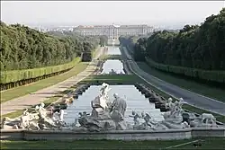 Frontale Farbfotografie einer rechteckigen Wasserfläche, die bis zum Hintergrund führt und an einem Schloss endet. Das Wasser wird durch zwei kleine Rasenflächen durchbrochen. An beiden Seiten sind Wege, Heckenreihen und Wälder. Im Vordergrund sind mehrere Skulpturen, die eine Szene darstellen.