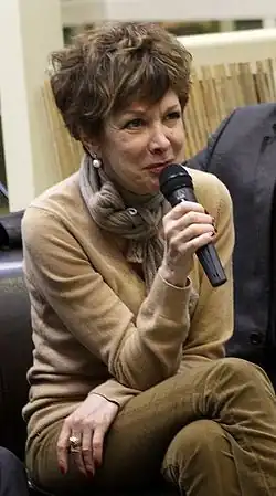 Catherine Laborde, 2010. Eine ältere Dame mit wuscheligen kurzen Haaren im braunen Strick-Pullover sitzt auf einer Couch und spricht in ein Handmikrophon.