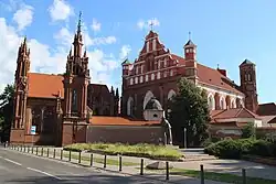 Seitliche Farbfotografie von zwei braunen Kirchen. Die linke Kirche hat zwei gotische Türme mit vielen Verzierungen. Die rechte Kirche hat vorne zwei kleine Ecktürme und ein großes, braun-weißes Giebelfeld mit einem gemalten Jesuskreuz. Hinter der Kirche ist ein breiter Turm im gotischen Stil.