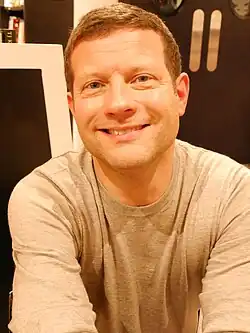 Dermot O’Leary sitzt vor einer dunkelbraunen Wand, neben der ein Bücherregal steht. Er trägt ein beiges T-Shirt, einen leichten Stoppelbart sowie seine hellbraunen Haare oben flach und an den Seiten bis zu den Ohren kurz geschnitten.